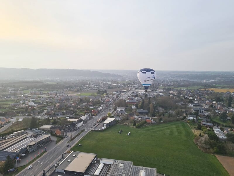 Montgolfière blanche avec logo survolant une zone urbaine et des champs verts à l'horizon.