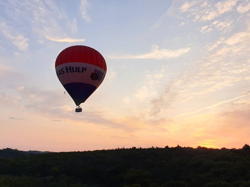 Montgolfière rouge et blanche flottant dans un ciel au coucher du soleil au-dessus d'une forêt.