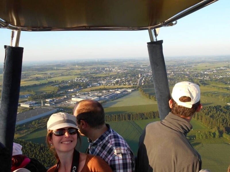 Trois personnes dans une nacelle de montgolfière souriantes, survolant une ville et des champs.