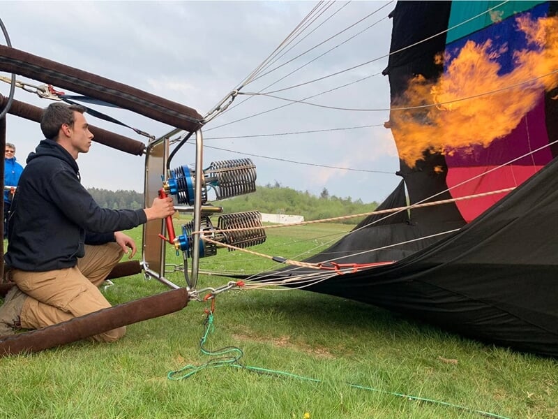Homme allumant le brûleur d'une montgolfière noire sur un terrain herbeux avant le décollage.