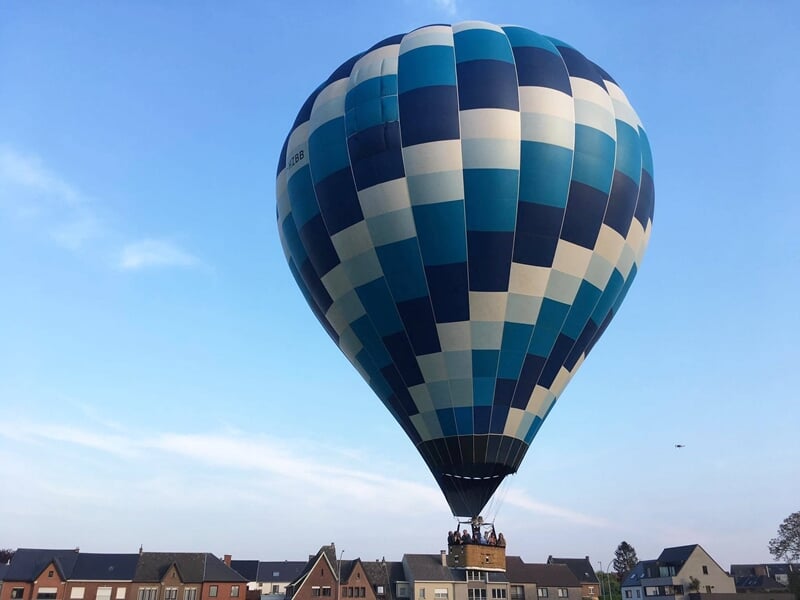 Montgolfière bleue et blanche en vol au-dessus de maisons et d'un paysage rural verdoyant.