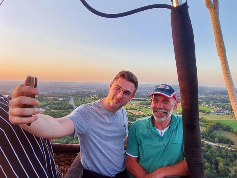 Deux hommes prenant un selfie dans une nacelle de montgolfière avec un paysage en arrière-plan.