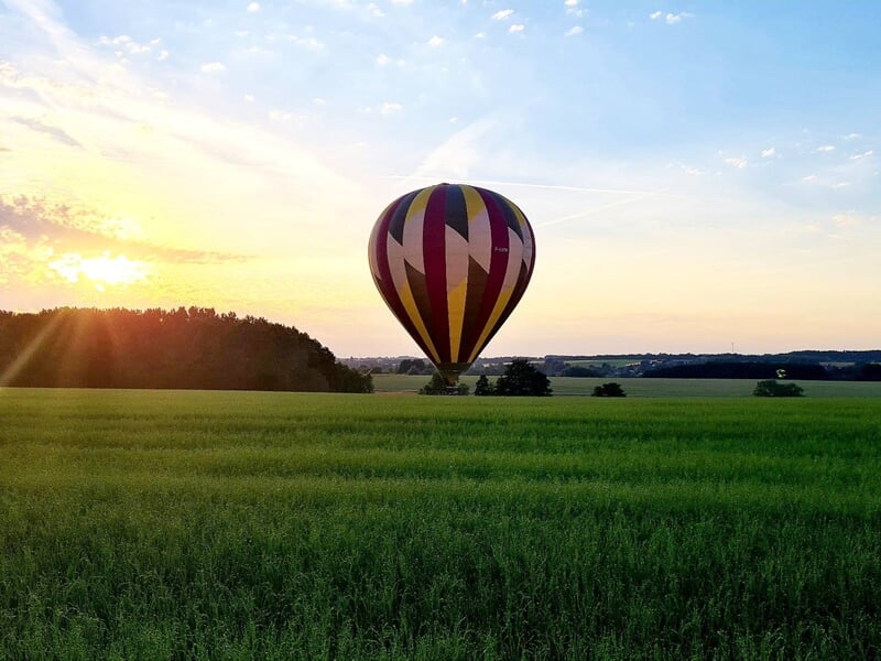 Montgolfière colorée au lever du soleil survolant un champ vert avec un ciel partiellement nuageux.
