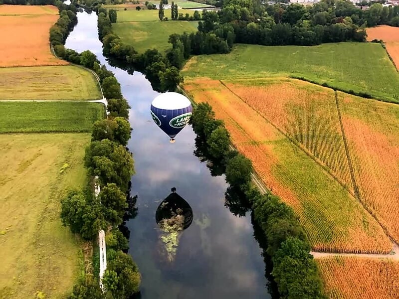 Montgolfière Land Rover volant au-dessus d'une rivière calme, reflet parfait dans l'eau, entourée de champs et d'arbres verdoyants.