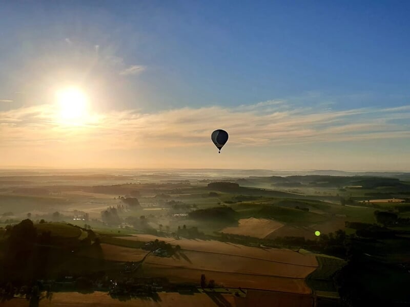 Montgolfière volant au lever du soleil au-dessus d'un paysage rural avec champs et collines verdoyantes.