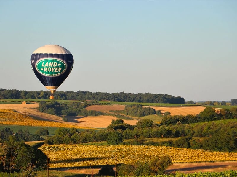 Montgolfière Land Rover en vol au-dessus de champs dorés et de collines boisées sous un ciel clair.
