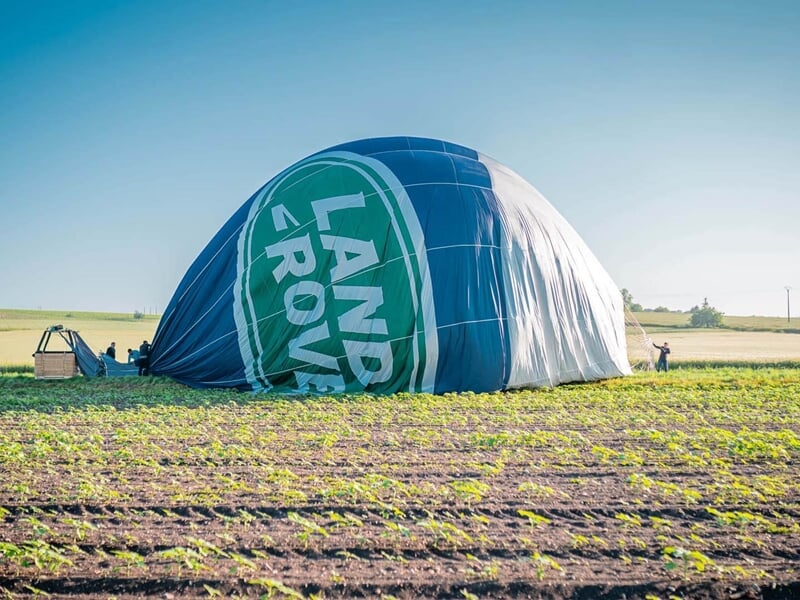 Montgolfière dégonflée avec logo Land Rover dans un champ agricole, entourée de verdure et ciel bleu.