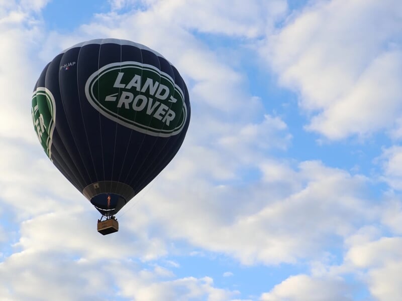 Montgolfière Land Rover en vol avec ciel bleu et nuages blancs, survolant un paysage verdoyant.