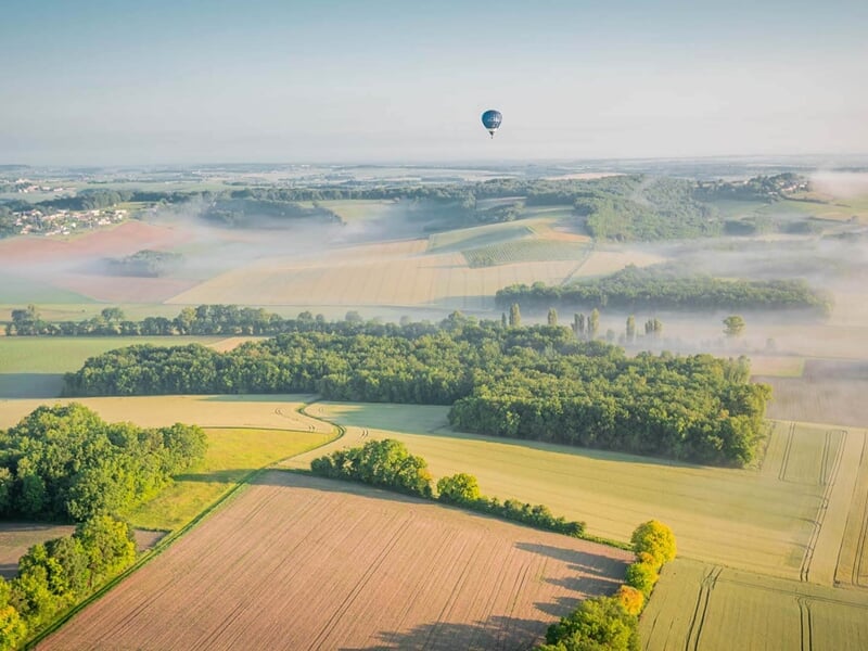 Montgolfière volant au-dessus de champs et forêts avec une légère brume matinale sous un ciel clair.