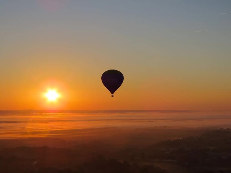 Montgolfière en vol au coucher du soleil, silhouette noire sur un ciel orange et paysage rural brumeux.