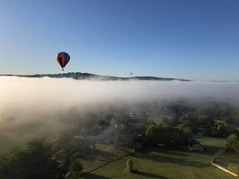 Vue aérienne d'un village avec maisons anciennes et collines boisées sous un ciel bleu clair.