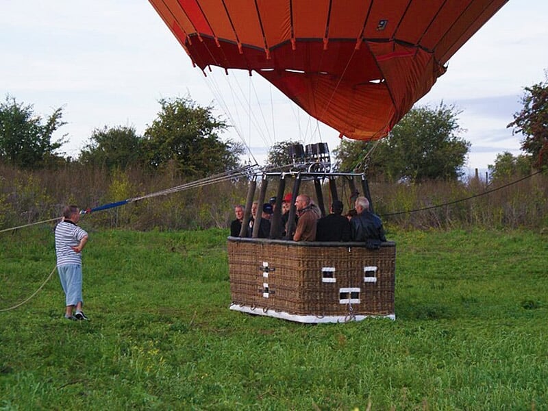 Groupe de personnes dans une nacelle de montgolfière au sol, entourée d'herbe et d'arbres en arrière-plan.