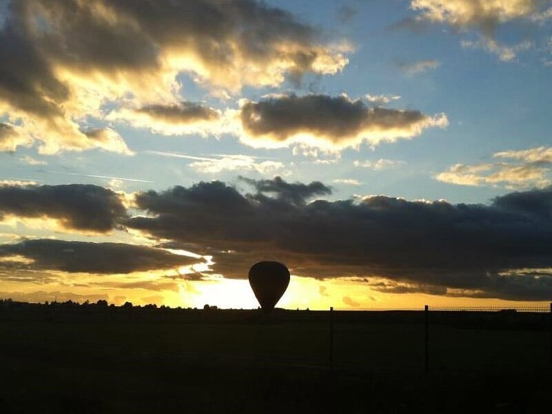 Montgolfière en silhouette au coucher du soleil, posée dans un champ avec un ciel nuageux coloré.