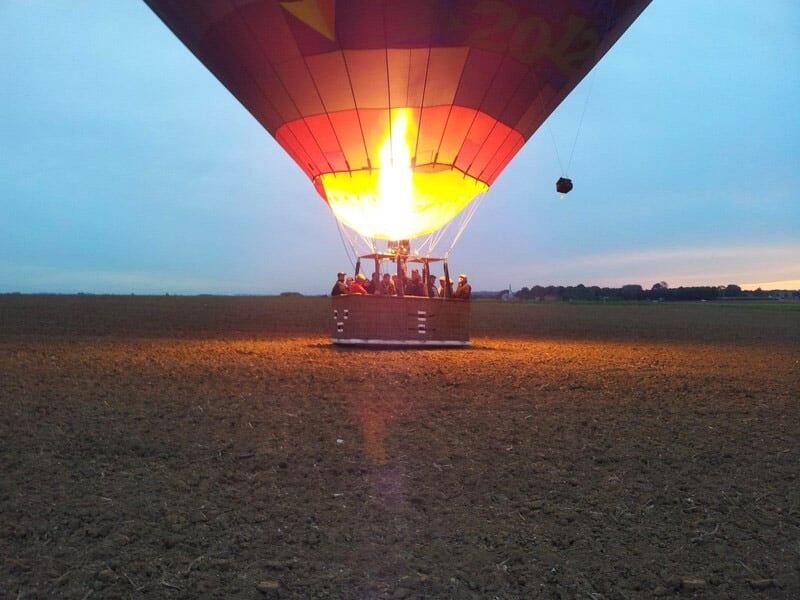 Montgolfière au sol avec la flamme du brûleur allumée, éclairant la nacelle dans un champ à la tombée du jour.