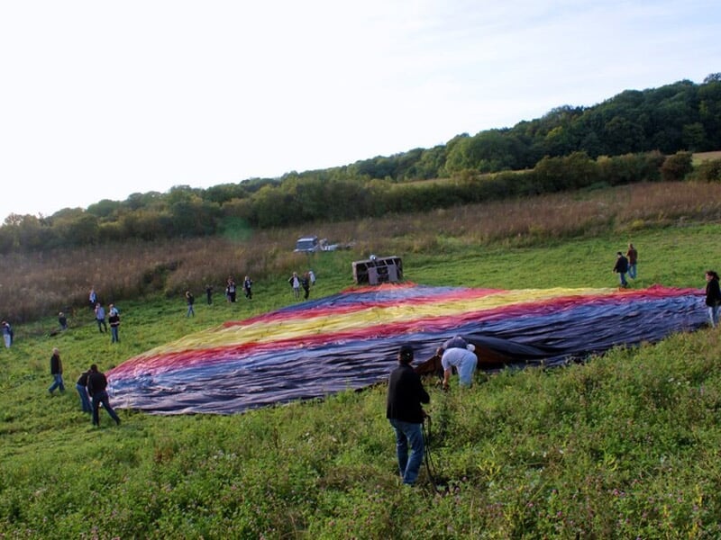 Groupe de personnes déployant la toile d'une montgolfière colorée dans un champ herbeux entouré d'arbres.