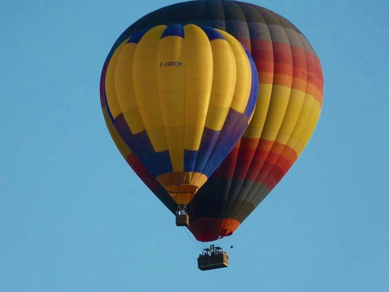 Deux montgolfières colorées en vol dans un ciel bleu clair, avec des passagers visibles dans les nacelles.