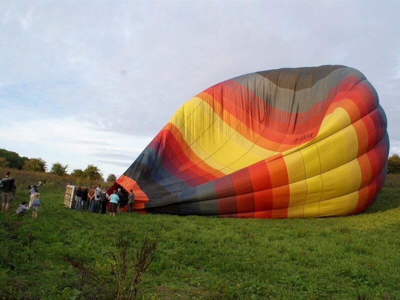 Montgolfière dégonflée au sol avec des personnes autour, dans un champ vert sous un ciel nuageux.