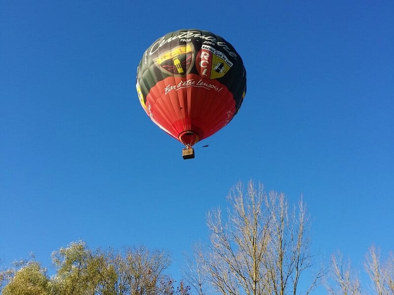 Montgolfière colorée en vol au-dessus d'un paysage rural avec des champs et des arbres sous un ciel clair.