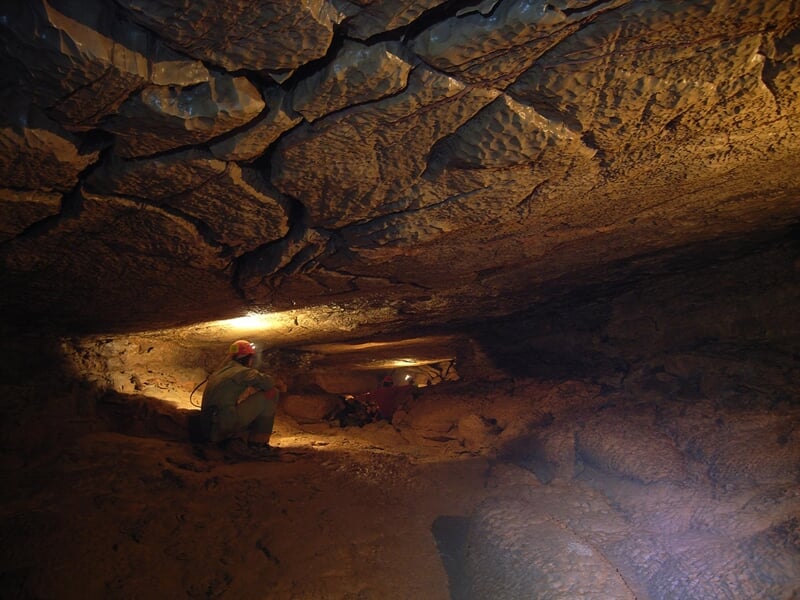 Spéléologue éclairé par une lampe frontale explore une grande cavité souterraine aux parois rocheuses et texturées, mettant en valeur les nuances de brun et les formations géologiques