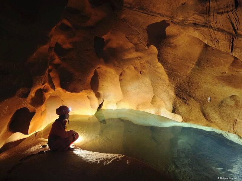 Explorateur assis à côté d'une flaque d'eau dans une grotte, éclairé doucement par sa lampe, révélant des parois lisses de couleur sable avec des ombres douces et des textures ondulées
