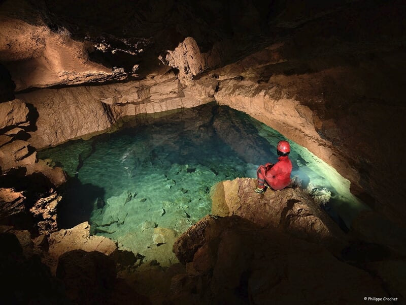 Spéléologue observant une étendue d'eau cristalline dans une caverne souterraine, éclairé par une lumière douce qui souligne les tons verts de l'eau et les formations rocheuses environnantes