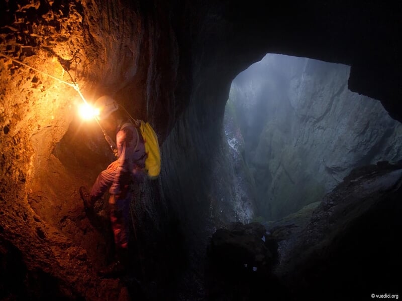 Spéléologue descendant une corde dans une grande ouverture de caverne avec une vue sur une vallée verdoyante visible à travers l'entrée, dans un environnement sombre éclairé par une lumière vive de la