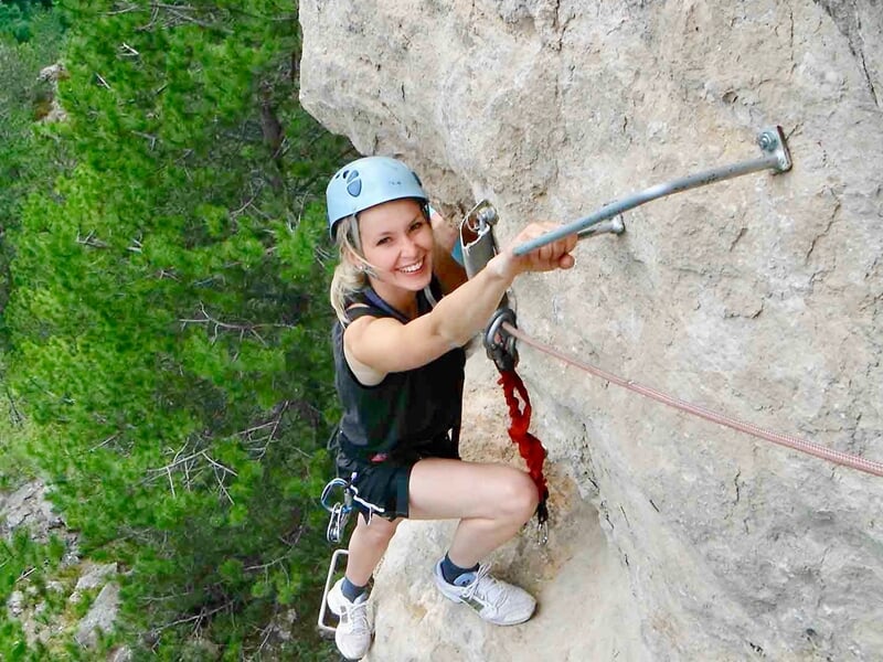 Femme souriante en casque escaladant une paroi rocheuse avec équipement de via ferrata en pleine nature.