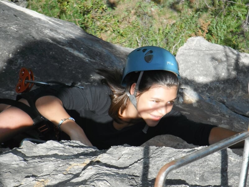 Femme concentrée en casque progressant sur une via ferrata rocheuse avec prises métalliques visibles.