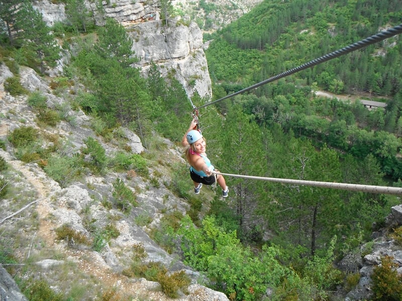 Personne suspendue à une tyrolienne au-dessus d'une vallée verdoyante avec forêt dense en arrière-plan.