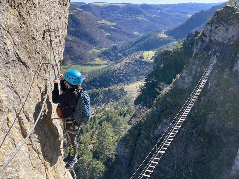 Enfant en casque escaladant une paroi rocheuse avec un pont suspendu visible dans un paysage montagneux.