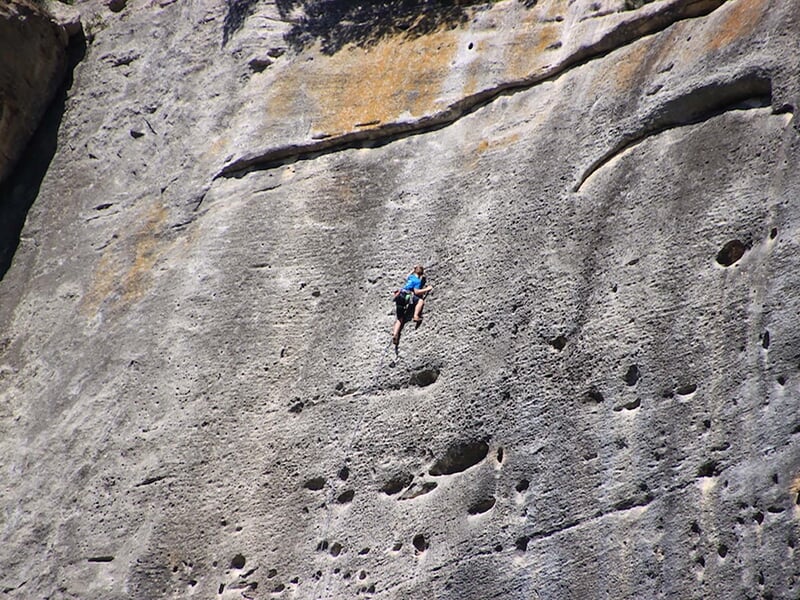 Grimpeur suspendu à une paroi rocheuse lisse avec équipement d'escalade sous un ciel bleu clair.