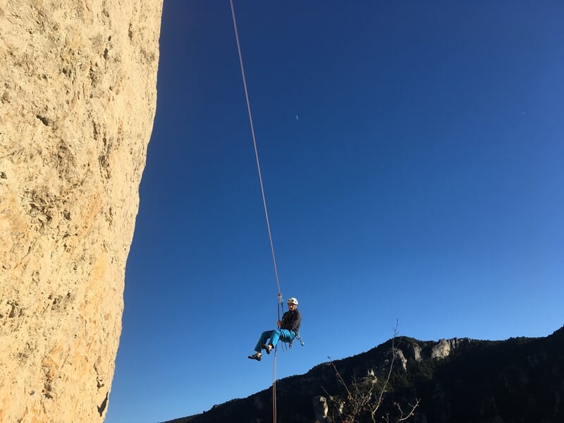 Personne en tenue d'escalade descendant en rappel le long d'une falaise rocheuse sous un ciel bleu.