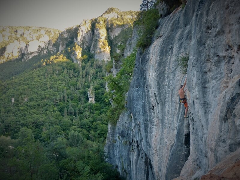 Grimpeur en falaise rocheuse avec paysage montagneux en arrière-plan sous une lumière naturelle.