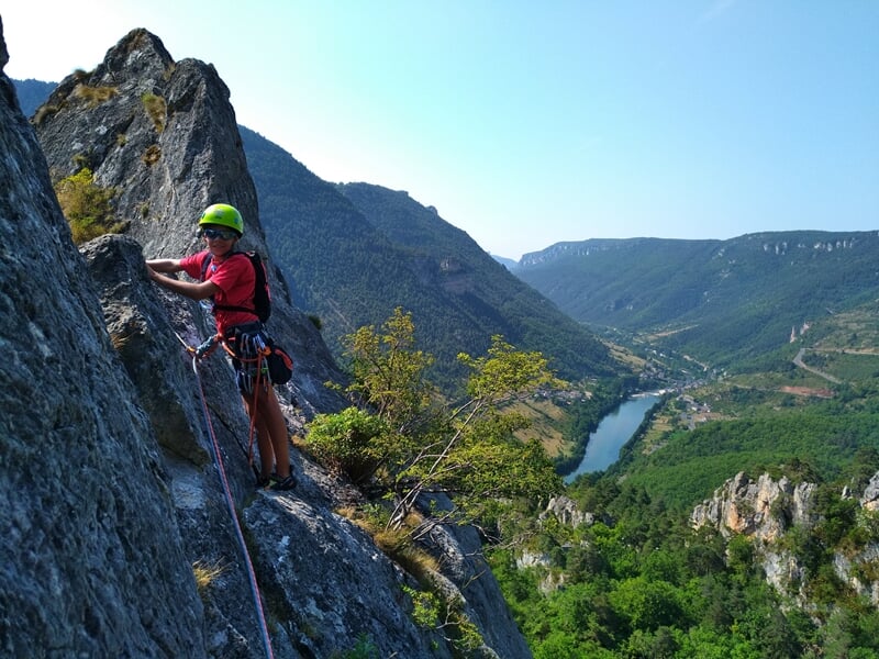 Escaladeur en casque progressant sur une paroi rocheuse abrupte avec vue sur vallée verdoyante.