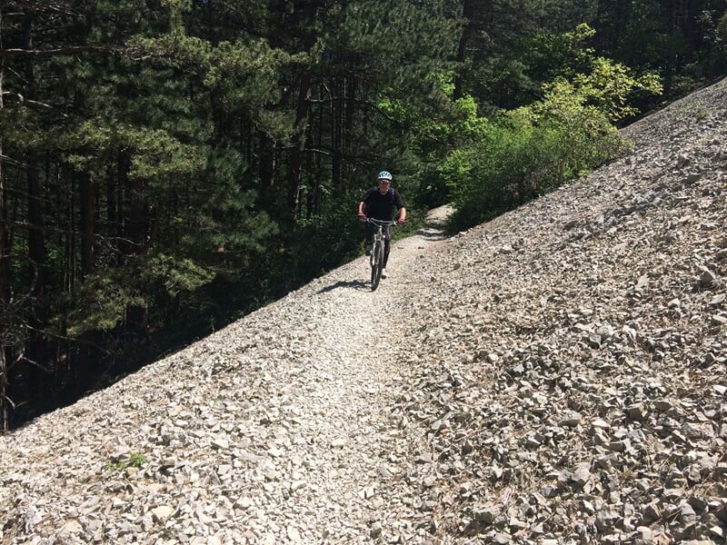 Cycliste en VTT roulant sur un chemin de terre caillouteux entouré de forêt dense et de rochers.