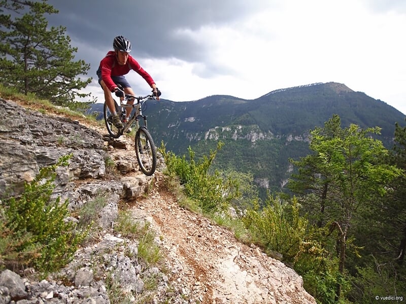 Personne en VTT sur un chemin de montagne rocheux avec vue panoramique sur un village en vallée.