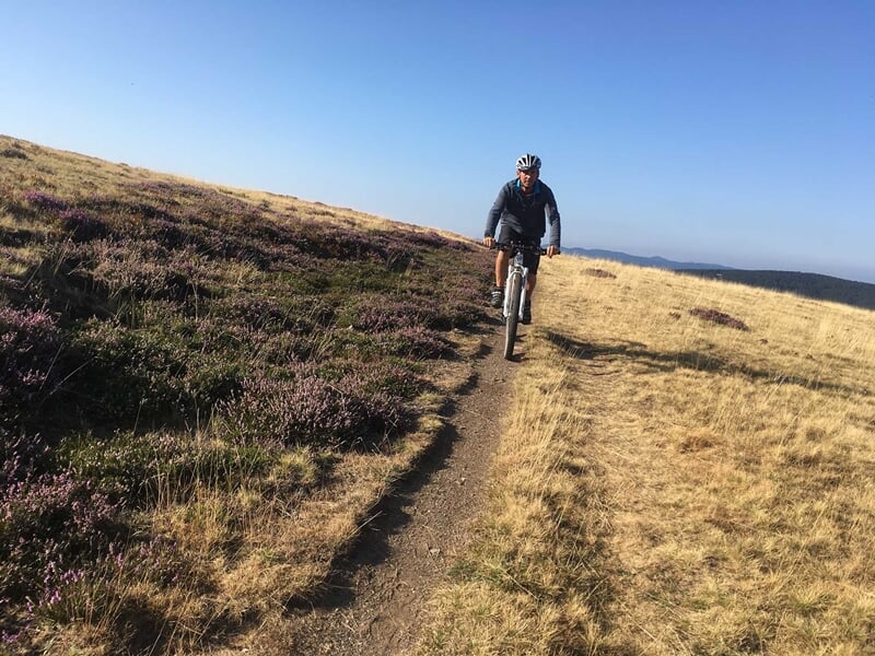 Cycliste en VTT sur un sentier étroit en montagne avec paysage de collines et végétation verte.