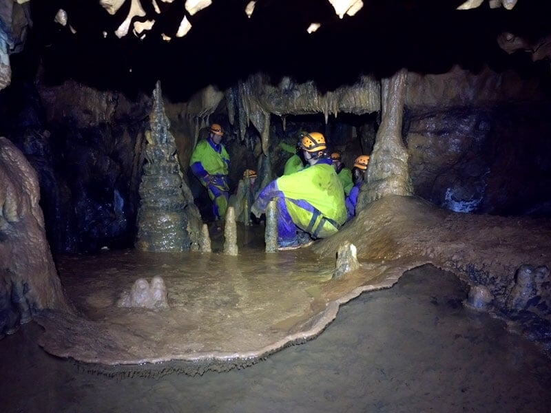 Groupe de spéléologues en équipement de protection, éclairés par leurs lampes frontales, discutant près d'une formation de stalagmite dans une grotte aux parois brunâtres et aux détails stalactitiques