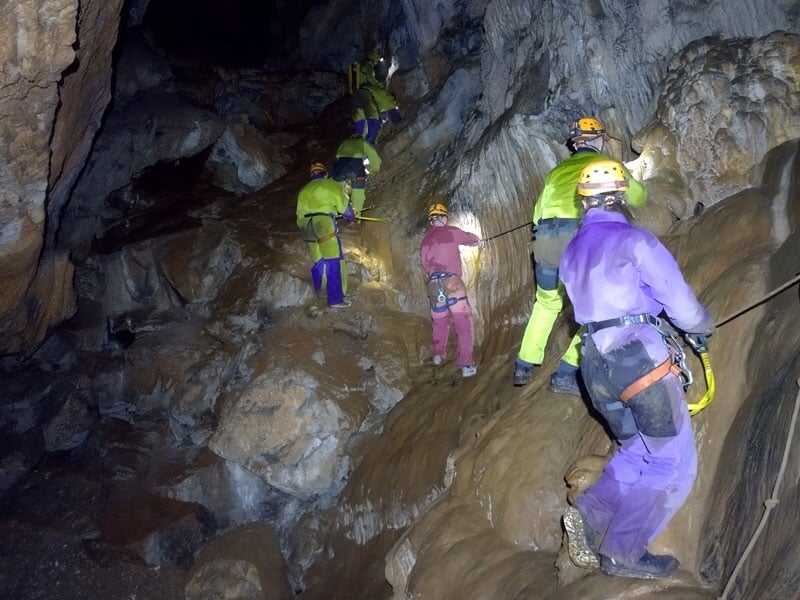 Spéléologues en tenue colorée équipés de harnais sécurisant leur progression sur une pente rocheuse et glissante dans une grande caverne, illustrant le travail d'équipe et l'aventure en exploration so