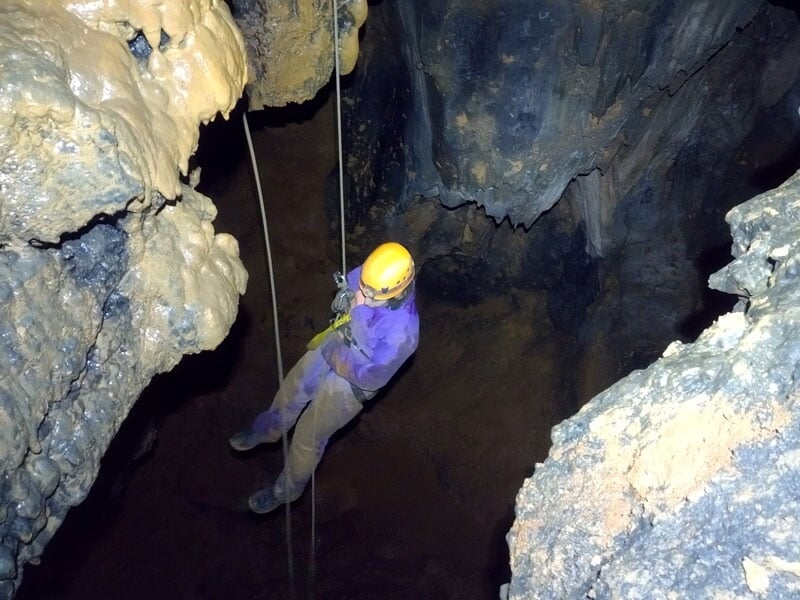 Spéléologue en descente sur corde, suspendu dans un grand vide souterrain avec des parois rocheuses sombres, mettant en évidence les techniques de descente en rappel et l'ampleur de la caverne exploré