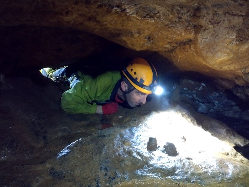 Spéléologue en combinaison verte et jaune, équipé d'un casque avec lampe frontale, progressant prudemment dans un passage étroit sous-terrain aux parois rocheuses humides et brillantes