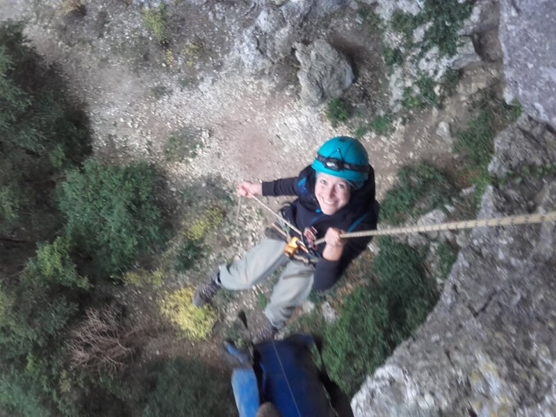 Personne en casque bleu descend en rappel sur une paroi rocheuse avec une forêt en arrière-plan.