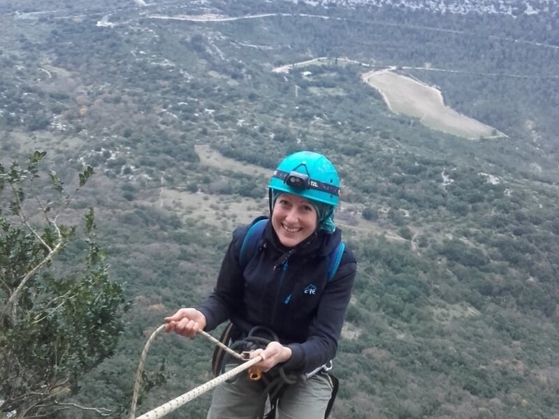 Femme en casque bleu souriante tient une corde d'escalade avec un paysage de collines en arrière-plan.
