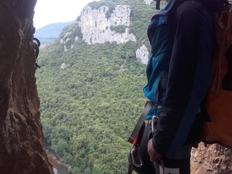 Personne équipée d'un sac à dos et casque regarde un paysage de falaises et forêt depuis une grotte.