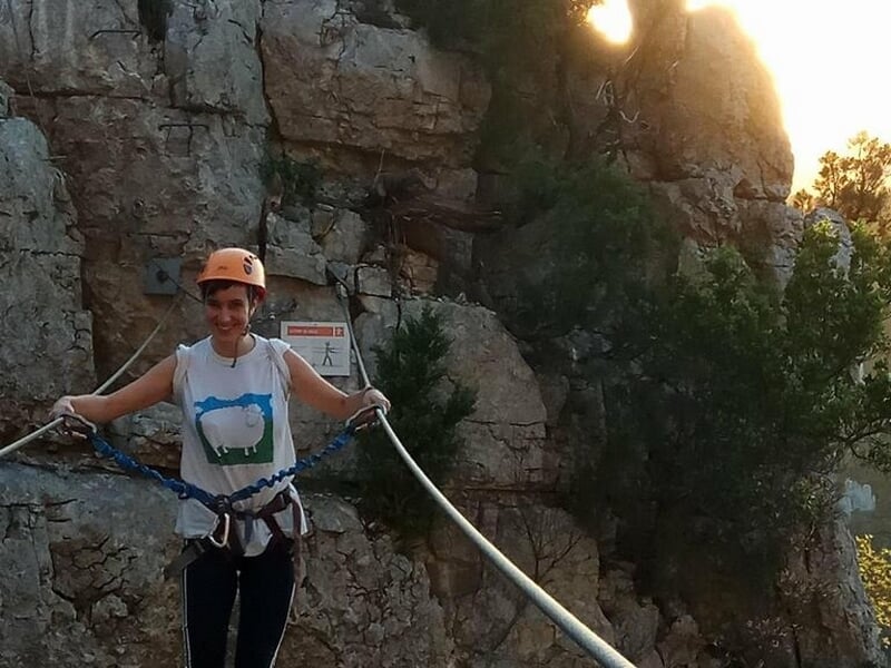 Femme en casque orange et harnais souriante tient une corde sur un sentier rocheux en via ferrata.