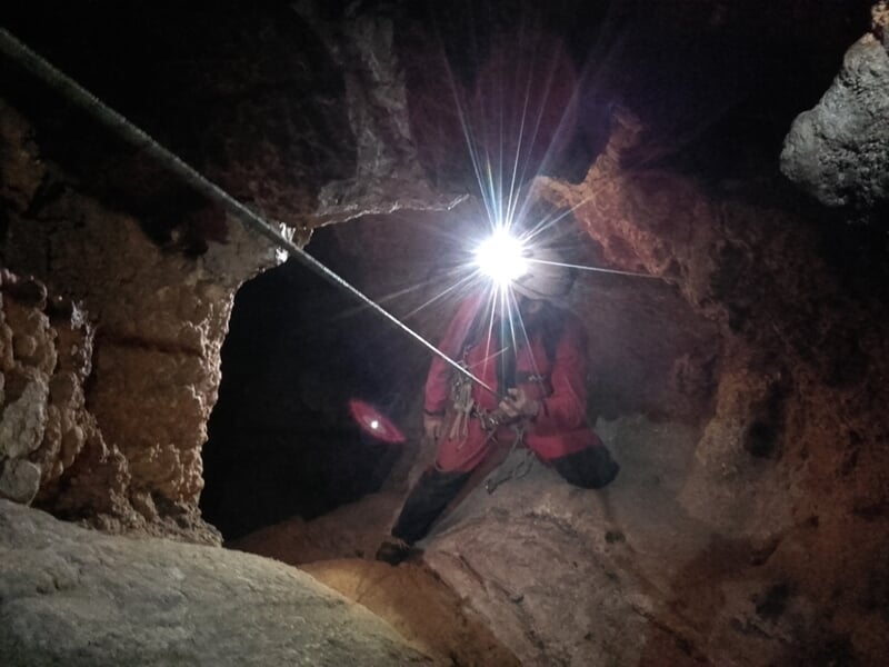 Spéléologue équipé d'un casque et lampe frontale descend en rappel dans une cavité rocheuse étroite.