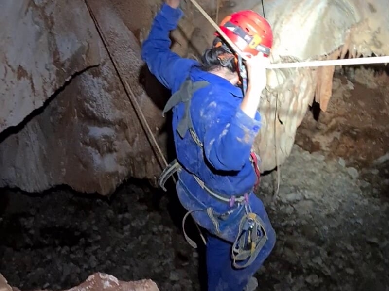 Personne en combinaison bleue et casque rouge descend une paroi rocheuse en spéléologie avec une corde.