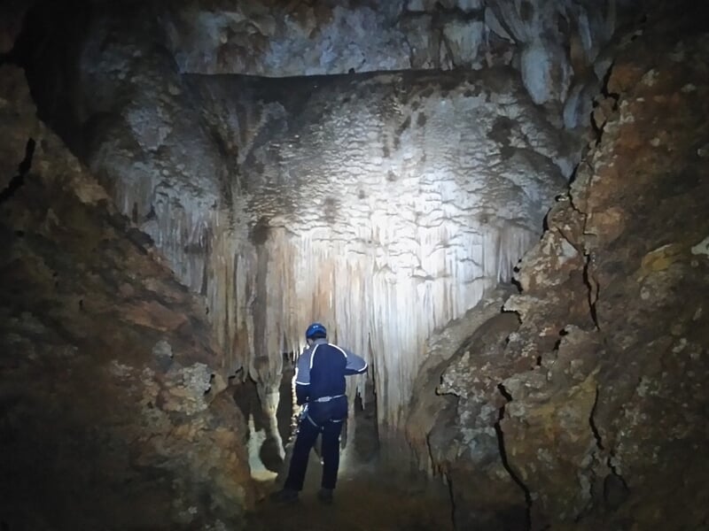 Spéléologue marche dans une grande salle souterraine avec des formations rocheuses et stalactites.