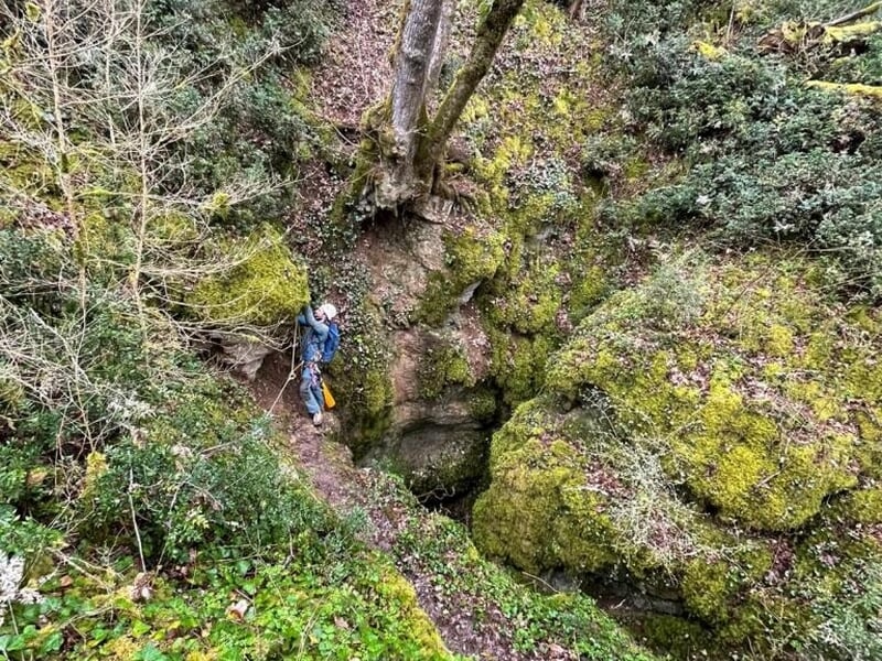 Sentier forestier verdoyant avec un spéléologue équipé d'un casque et sac à dos près d'une cavité.