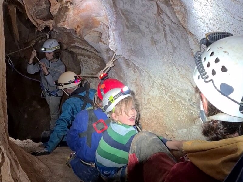 Groupe de spéléologues souriants équipés de casques et harnais progressent dans une grotte étroite.